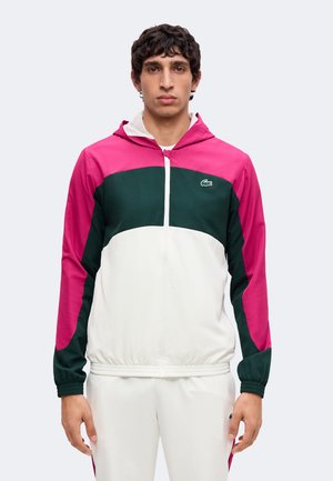 Mann mit Blick nach vorne, trägt eine color-block Jacke mit rosa, dunkelgrünen und weißen Bereichen und passende weiße Hose vor schlichtem Hintergrund.