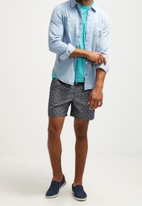 Light blue denim shirt over a teal t-shirt, paired with dark shorts featuring white polka dots. Dark blue slip-on shoes complete the look.