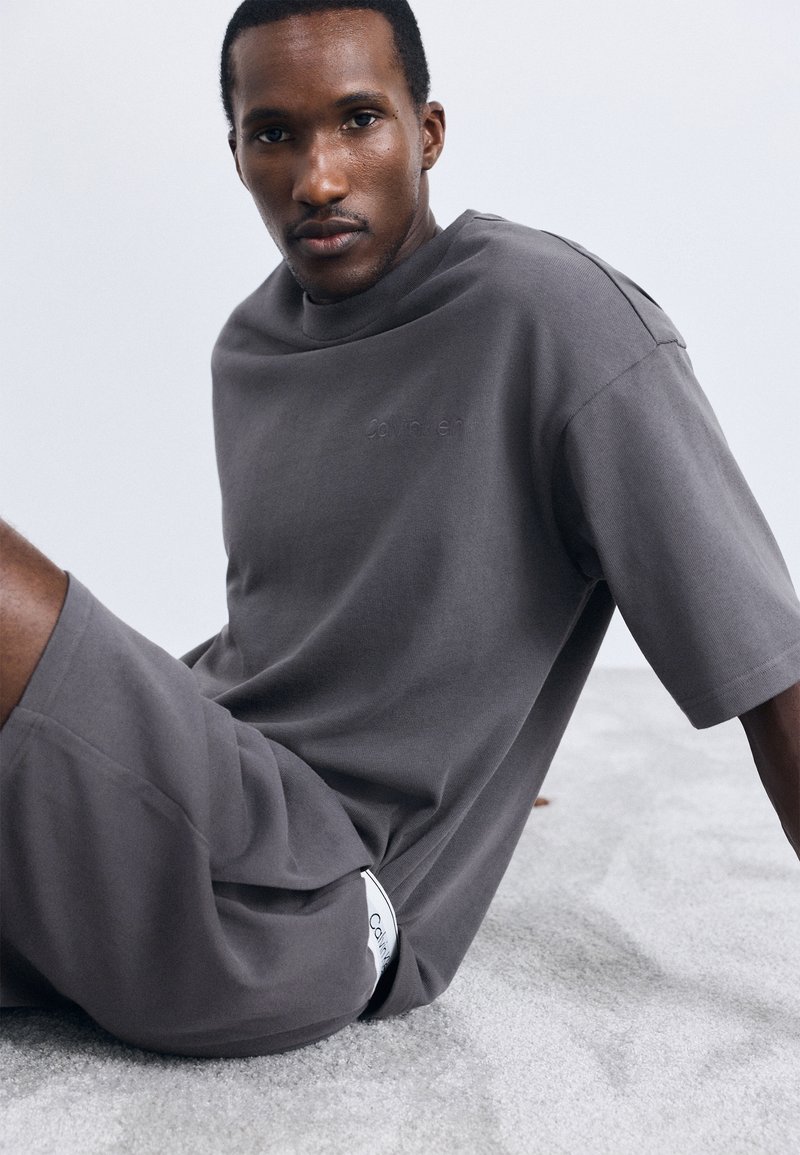 Tricou din bumbac gri, cu mâneci scurte și o croială lejeră. Prezintă branding subtil și un etichetă pe lateral, asortat cu pantaloni scurți. Material texturat.