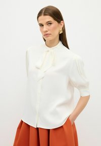 Camicia bianca con colletto a fiocco, maniche a palloncino corte e texture liscia, abbinata a una gonna color ruggine.