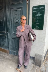 Oversized brown suit with a single-breasted jacket and wide-leg trousers. Carrying a black and white handbag. Grey door and plaque backdrop.