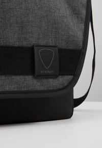 Borsa a tracolla grigia con corpo in tessuto texturizzato, rifiniture nere e patch in gomma con logo. Presenta una tracolla regolabile e un design elegante e minimalista.