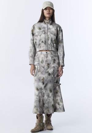 Femme portant une casquette beige, une veste courte tie-dye à poches avant, une jupe longue assortie avec poches cargo, et des bottines camel à franges.