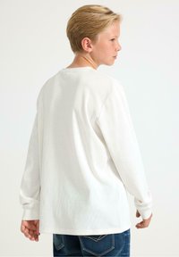 Witte katoenen blouse met lange mouwen, een ronde halslijn en ribbeltextuur, gedragen met blauwe spijkerbroeken, van achteren weergegeven.