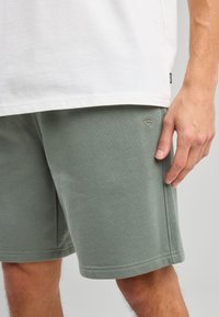 Green cotton shorts with a textured finish and side pockets. Features a small embroidered logo near the hem. Worn with a white t-shirt.