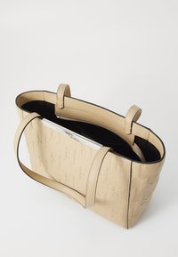 Beige tote veske med vevde mønster, med svart innvendig, to håndtak og en glidelåslomme. Glatt tekstur med et minimalistisk design.