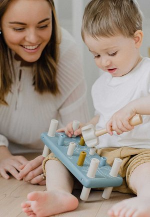 Wooden hammer toy in pastel blue, with six colorful pegs and white legs. Soft, rounded edges; child uses a wooden hammer to play.