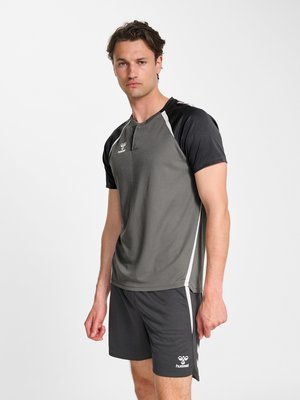 Mężczyzna w szarej i czarnej sportowej koszulce i szortach stoi na jednolitym białym tle z neutralnym wyrazem twarzy.