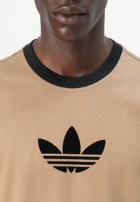 Bežinis marškinėliai su juodu apykaklės kraštu ir juodu Adidas logotipu su trimis juostelėmis po lapo emblema, pagaminti iš lygaus audinio.