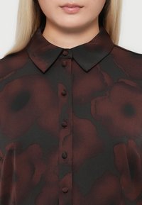 Gros plan sur la poitrine d'une femme portant un chemisier transparent foncé avec de grands motifs floraux bordeaux et des boutons recouverts de tissu sur le devant.