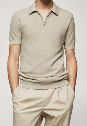 Mies, jolla on beige lyhythihainen neulottu polopaita vetoketjukauluksella ja vaaleanbeiget laskostetut housut, kädet taskuissa.