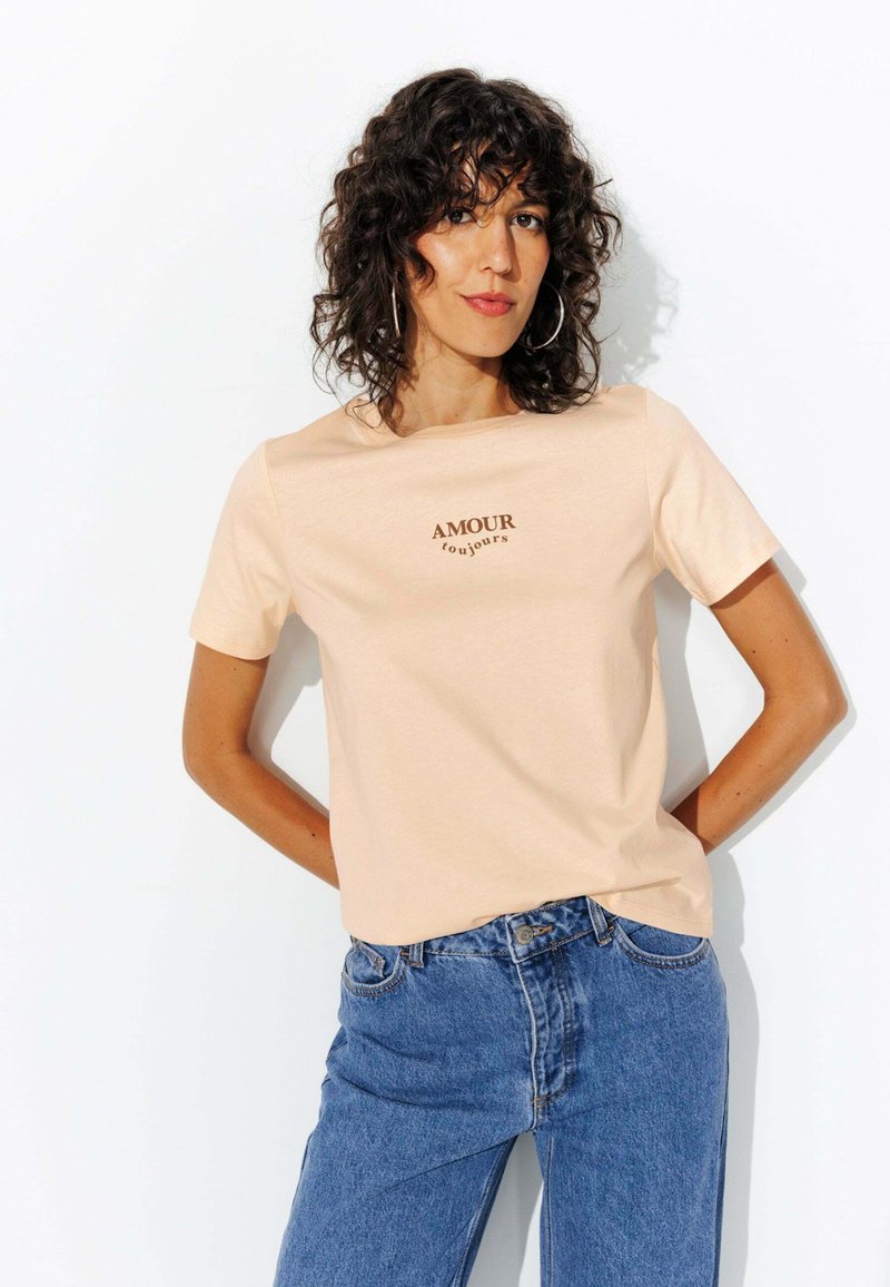 T-shirt en coton couleur pêche à manches courtes, arborant "AMOUR toujours" en écriture brun foncé sur la poitrine, assorti à un jean en denim bleu.