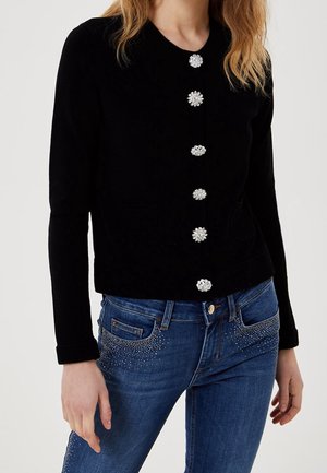 Femme portant un haut noir à manches longues avec six boutons décoratifs ornés de pierres précieuses, associé à un jean bleu avec de petits détails en strass.