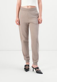Vrouw in beige joggingsbroek en zwarte high-heeled mules met spitse neus, gespsluiting en oogdetail.