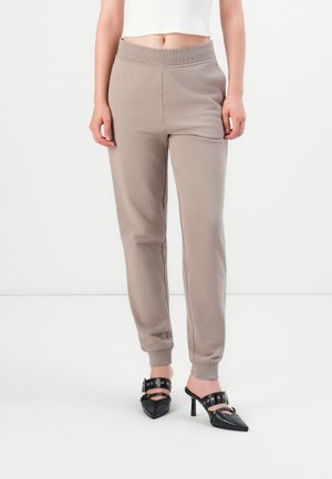 Kvinne iført beige joggebukser og svarte høyhælte spisse mules med spenne og hulldetaljer.
