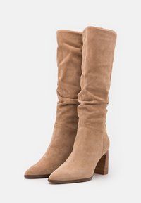 Bottes hauteur genou en daim beige avec un design décontracté, bout pointu et un talon bloc en bois texturé. Texture lisse sans motifs visibles.