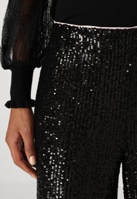 Black sequined pants with a textured surface, featuring a fitted design and a delicate ruffled trim at the waistband.