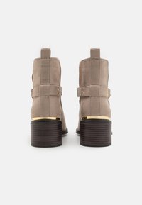 Beige suede ankle boots with a gold accent on the heel, featuring a block heel and side straps. Back view highlights stitching details.
