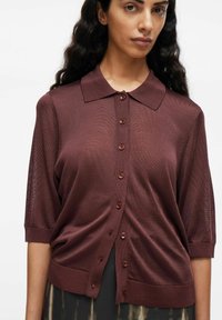Donna con capelli lunghi e scuri che indossa un cardigan corto a maniche corte marrone con colletto su uno sfondo bianco uniforme.