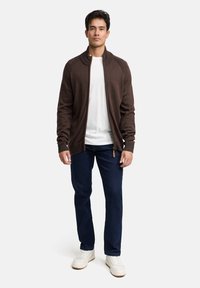 Brun sweatertop med lynlås og ribbet detalje ved skuldrene, båret over en hvid skjorte, kombineret med mørkeblå jeans og hvide sneakers.