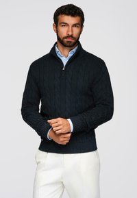 Homme avec une barbe portant un pull à col montant noir en tricot torsadé sur une chemise bleu clair et un pantalon blanc, se tenant devant un fond uni.