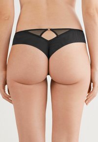 Zwarte stringonderbroek met een sheer mesh paneel en een kleine ronde accent achteraan, wat de minimalistische uitstraling benadrukt.