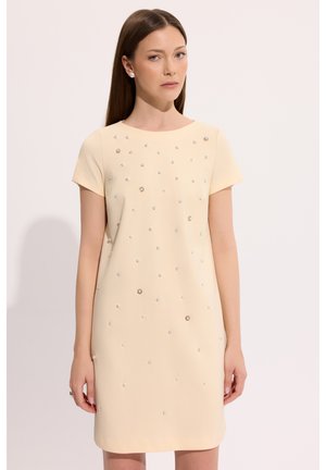 Kobieta o prostych brązowych włosach, ubrana w beżową sukienkę z krótkim rękawem, ozdobioną rozsianymi małymi kryształkami, stojąca na jednolitym białym tle.
