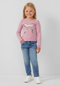 Chemise rose clair à manches longues avec un motif de nœud argenté, associée à un jean slim bleu et des baskets blanches. Texture de tissu lisse.