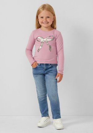 Hellrosa Langarmhemd mit grafischem silbernen Schleifenmotiv, kombiniert mit blauen Slim-Fit-Jeans und weißen Sneakers. Glatte Stofftextur.