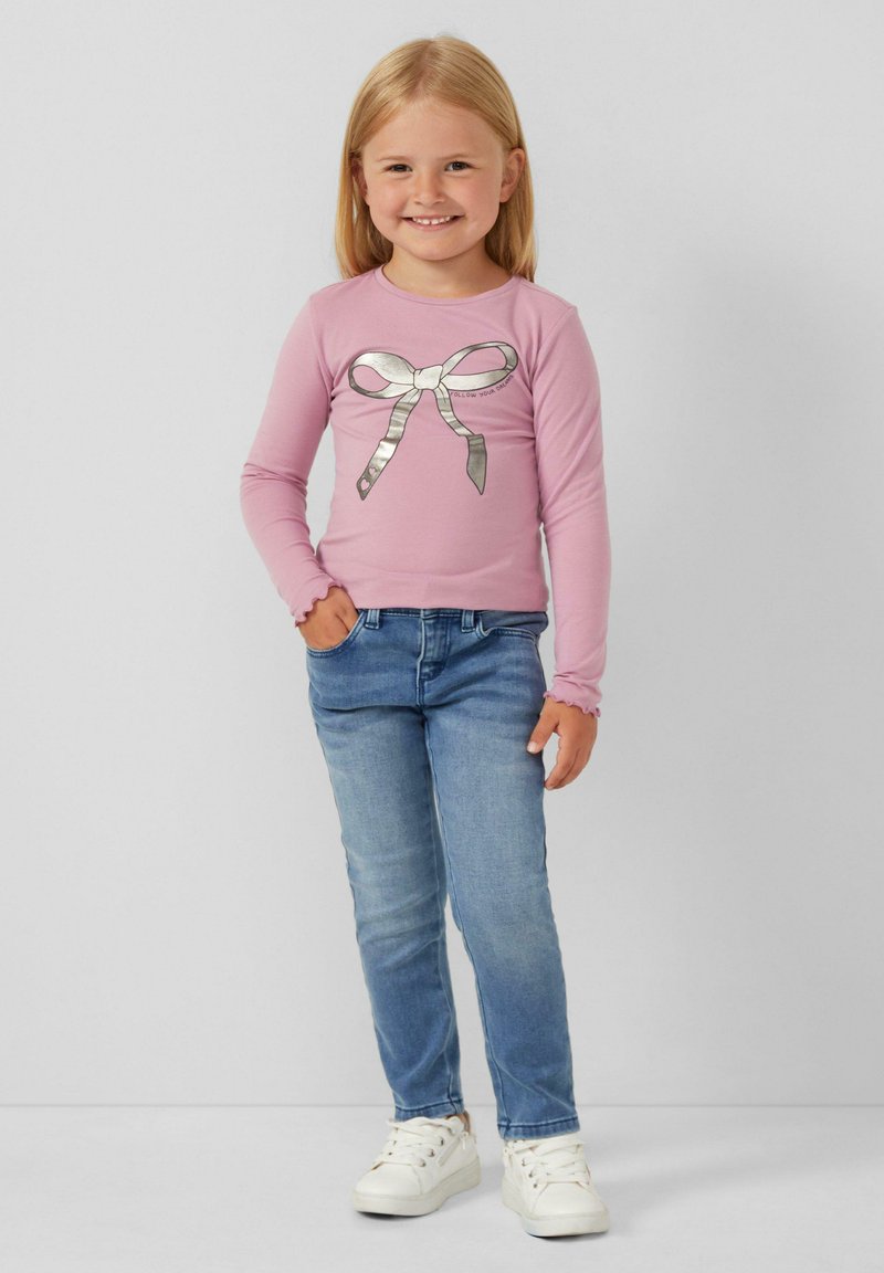 Chemise rose clair à manches longues avec un motif de nœud argenté, associée à un jean slim bleu et des baskets blanches. Texture de tissu lisse.