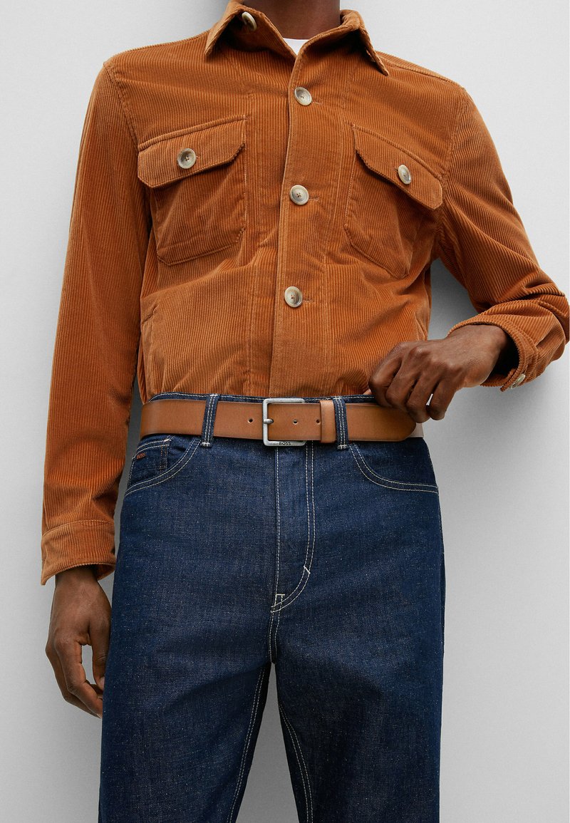 Oranges Corduroy-Hemd mit zwei Brusttaschen und Knopfverschlüssen, kombiniert mit dunkelblauen Jeans und einem braunen Ledergürtel mit einer silbernen Schnalle.