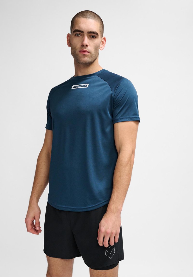Ung mand iført en mørkeblå Hummel sportst-shirt og sorte shorts står foran en ensfarvet lys baggrund.