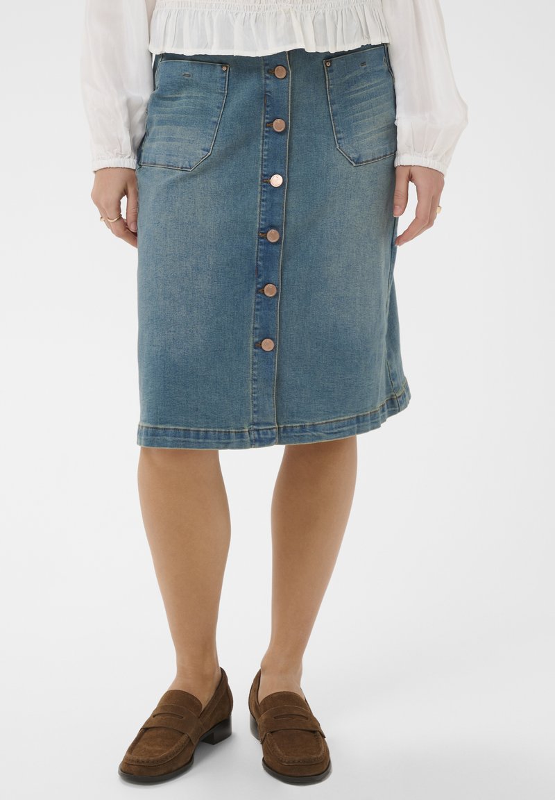 Ženska, ki nosi knee-length jeans krilo na gumbe z žepi spredaj, obuta v rjave loaferje in oblečeno belo majico z dolgimi rokavi.