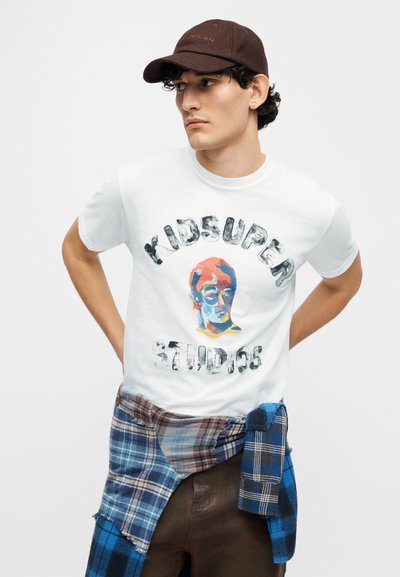 Λευκό βαμβακερό μπλουζάκι με πολύχρωμο γραφικό ενός προσώπου και τύπωμα «KIDSUPER STUDIOS». Καφέ καπέλο και καρό πουκάμισο δεμένο γύρω από τη μέση.