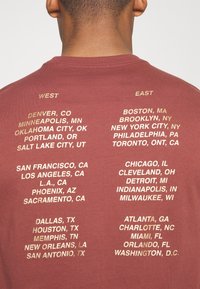 Punainen t-paita, jossa on kultaisia tekstejä, joissa on listattu kaupunkien nimiä, ja jotka on jaettu "Länsi"- ja "Itä"-osioihin, pyöreällä kauluksella ja lyhyillä hihoilla.