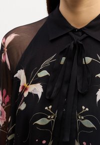 Blouse noire transparente avec des motifs floraux, un nœud noué au col et un col noir uni, montrant le haut de la poitrine et la zone des épaules.