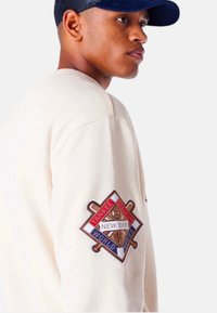 Sudadera de manga larga color crema con un parche bordado en marrón y rojo que presenta "Yankee Stadium" y "World Series" en el brazo izquierdo.