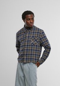 Férfi kék és zöld kockás ingben, mintás mellzsebbel, áll, az egyik keze világoskék farmerzsebben van.