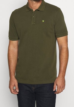 Man wearing an olive green short-sleeve polo shirt with a small green logo on the chest, paired with dark blue jeans.