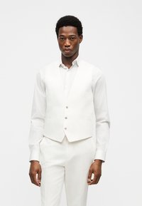 Άνδρας που φοράει άσπρο πουκάμισο, λευκό γιλέκο με τρία κουμπιά και λευκό παντελόνι απέναντι σε απλό φωτεινό φόντο.