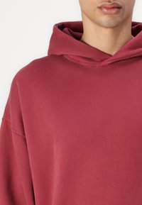 Hanorac bordeaux cu o croială relaxată, glugă mare și textură moale. Prezintă umeri căzuți și detalii de cusătură minime.