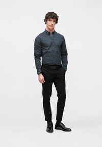 Mies seisoo käsineen taskuissa, pukeutuneena tummakuosiseen kauluspaitaan, mustiin housuihin, mustaan vyöhön, kelloon ja mustiin juhlakengiin valkoisella taustalla.