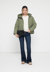 Giacca puffer verde oliva con cappuccio, abbinata a un maglione bianco, jeans flare in denim scuro e pochette nera. Scarpe beige con dettagli.