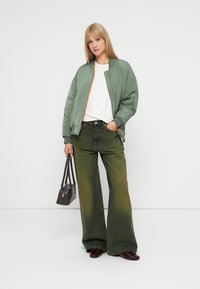 Μαξιλαρωτό μπόμμπερ τζάκετ σε ελιά με πορτοκαλί λεπτομέρεια στο φερμουάρ, συνδυασμένο με φαρδύ παντελόνι σε ξεθωριασμένο πράσινο και μια μικρή μαύρη τσάντα.