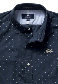 Granatowa koszula na guziki z drobnym żółtym wzorem w romby, haftowane logo La Martina z dwoma graczami polo na piersi, widoczna metka z rozmiarem M.