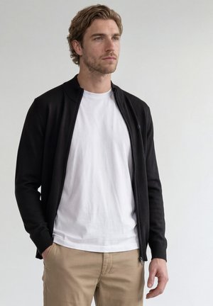 Hombre con cabello corto y castaño que lleva una camiseta blanca, chaqueta negra con cremallera y pantalones beige, de pie con una mano en el bolsillo frente a un fondo liso.