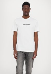 Fehér, rövid ujjú pamut póló, amelyen a középen fekete "ARMANI EXCHANGE" logó található. Szürke farmerrel viselve.