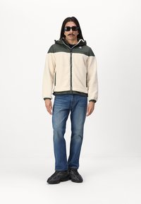 Giacca in pile di colore crema e verde scuro, con tessuto zip frontale, cappuccio regolabile e tasche laterali. Indossata con jeans blu e scarpe da ginnastica nere.