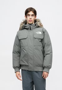 Graue isolierte Parka mit einer fellgefütterten Kapuze, Reißverschluss vorne, zwei Brusttaschen und gerippten Bündchen. Glattes Gewebe mit matter Oberfläche.