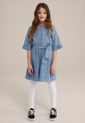 Abito di denim blu con maniche corte arricciate, caratterizzato da una chiusura a bottoni sul davanti, vita con cintura e gonna svasata. Abbinato a leggins bianchi e sneaker nere.
